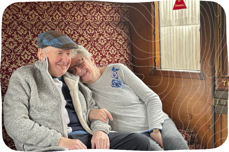 Mature man and his wife sitting inside a steam train. They are leaning against each other and smiling.