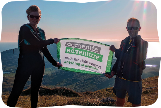 Two people holding a Dementia Adventure flag on top of a peak.