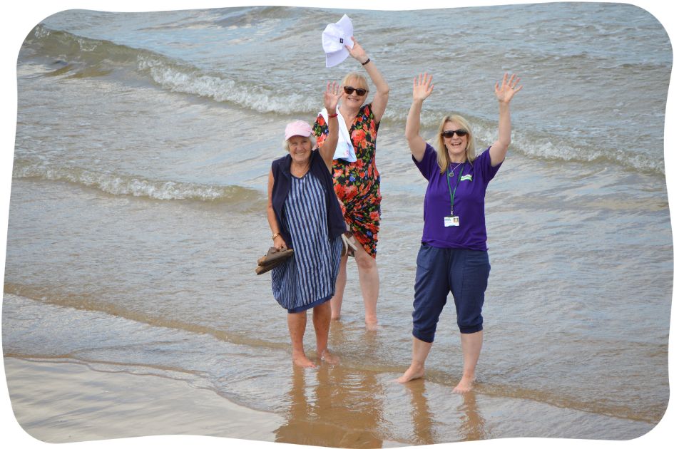 Three women standing barefoot in shallow waves on the beach, with their arms in the air waving.