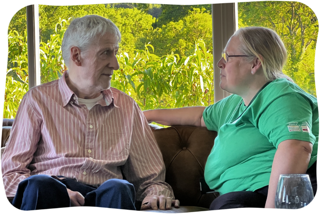 A man and woman sit on the sofa together, with a window looking out onto trees in the background.