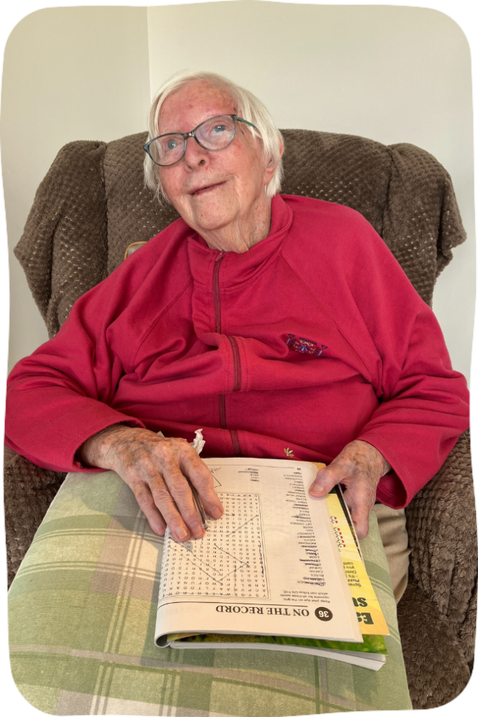 An elderly woman in a red fleece, sitting in an armchair and holding a pen and a booklet containing a worsearch puzzle.