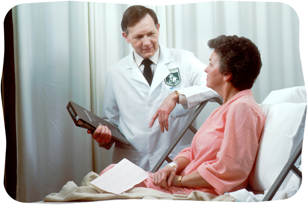 A male doctor in a white coat speaks to a female patient sitting up in a hospital bed.